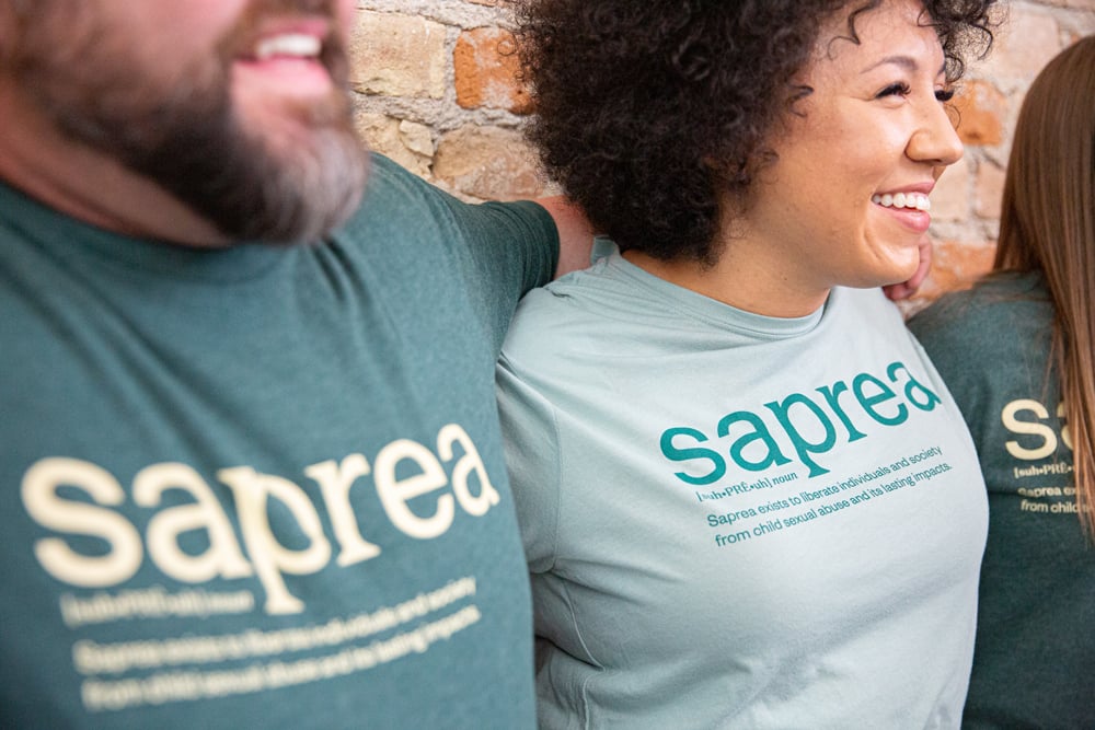 Close-up of three people wearing branded Saprea t-shirts, photographed against a brick wall